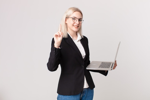 Blonde mooie vrouw die lacht en er vriendelijk uitziet, nummer één toont en een laptop vasthoudt