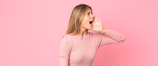 Blonde mooie vrouw die luid en boos schreeuwt om ruimte aan de zijkant te kopiëren, met de hand naast de mond