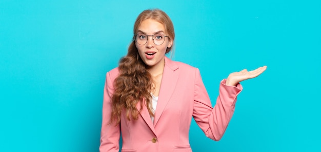 Blonde, mooie vrouw die verbaasd en geschokt kijkt, met open mond een voorwerp vasthoudt met een open hand op de zijkant