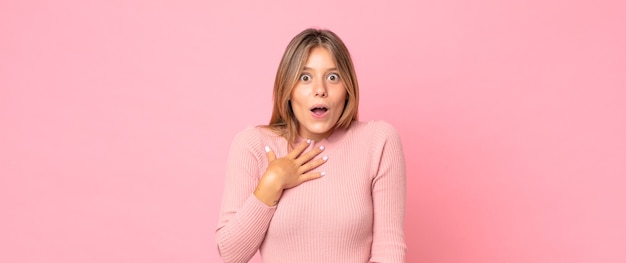 Blonde mooie vrouw die zich geschokt en verrast voelt, glimlacht, hand ter harte neemt, blij is om degene te zijn of dankbaarheid toont