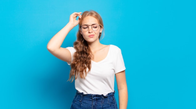 Blonde, mooie vrouw die zich verbaasd en verward voelt, haar hoofd krabt en naar de zijkant kijkt