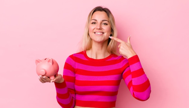 Blonde mooie vrouw glimlachend vol vertrouwen wijzend naar eigen brede glimlach spaarvarken concept