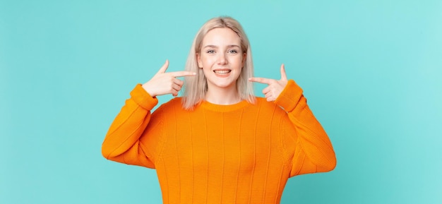 Blonde mooie vrouw glimlachend vol vertrouwen wijzend naar eigen brede glimlach