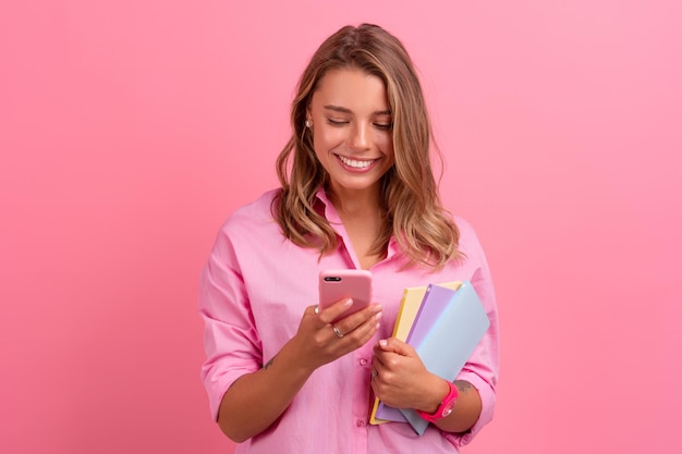 Blonde mooie vrouw in roze shirt glimlachend met notitieboekjes vast te houden en smartphone te gebruiken