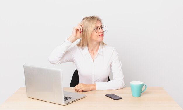 Blonde mooie vrouw jonge blonde vrouw die zich verbaasd en verward voelt en aan het hoofd krabt en met een laptop werkt