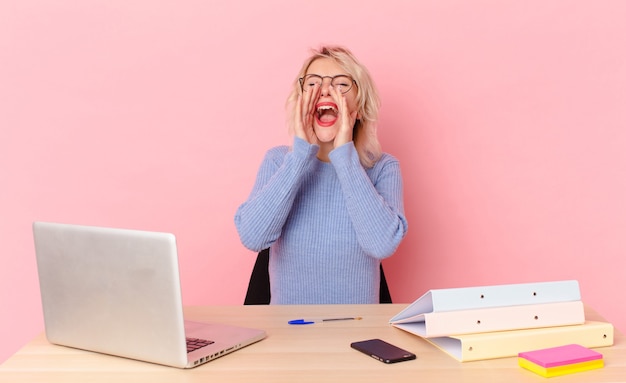 Blonde mooie vrouw jonge mooie vrouw die zich gelukkig voelt, een grote schreeuw geeft met de handen naast de mond. werkruimte bureau concept