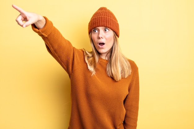 Blonde mooie vrouw voelt zich geschokt en verrast, wijst en kijkt vol ontzag naar boven met een verbaasde blik met open mond