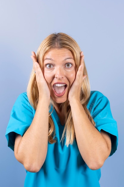 Blonde nurse in blue scrubs scared with her hands on her face