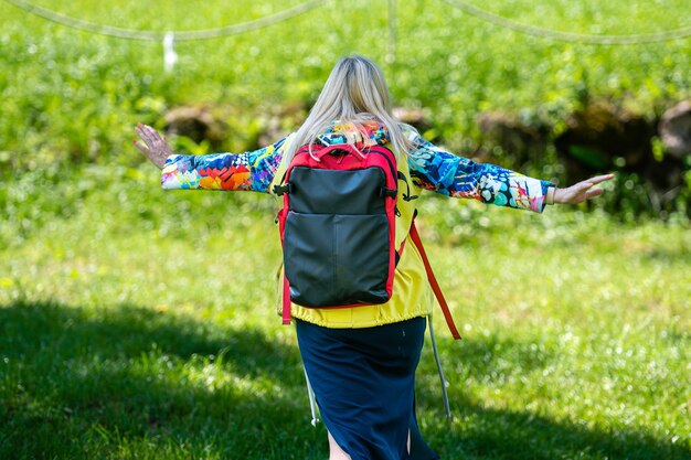 Foto blonde oudere vrouw in heldere kleren met een rugzak in het park op een zonnige dag achteruitkijk