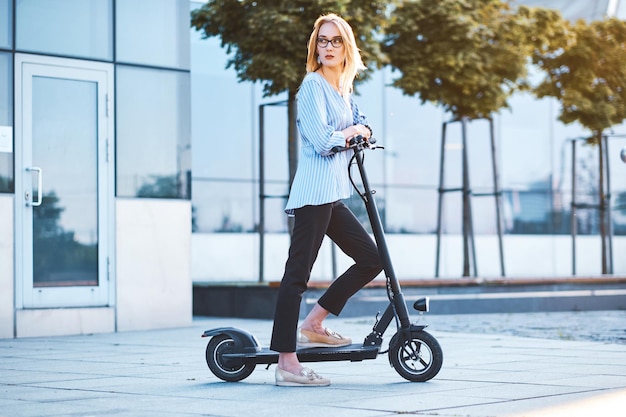 Blonde peinzende student met een bril rijdt met haar nieuwe effen zwarte electro-scooter naar de universiteit.