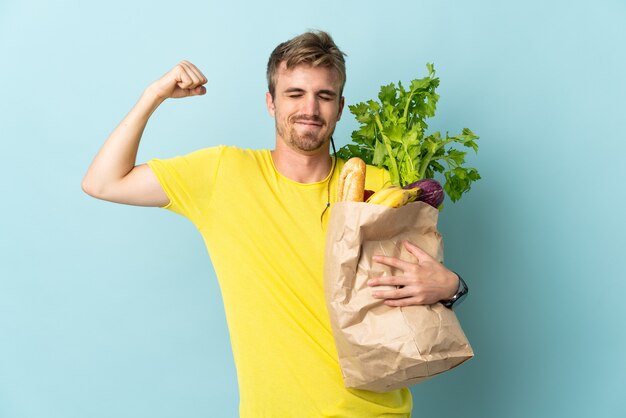 Blonde Person taking a bag of takeaway food isolated on blue background doing strong gesture