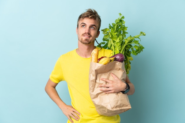 Persona bionda prendendo un sacchetto di cibo da asporto isolato su sfondo blu pensando un'idea mentre guarda in alto