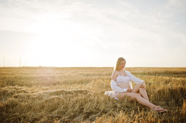 Blonde pregnant woman in wreath field at white underwear clothes on sunset