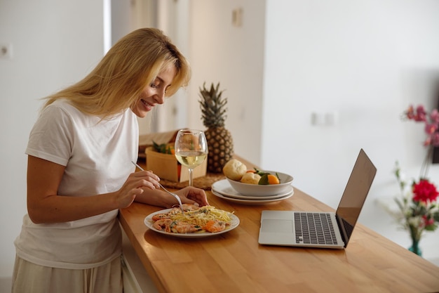 ラップトップでキッチンでエビとパスタを食べる金髪のきれいな女性昼食のためのワインのグラス