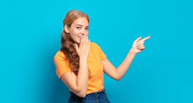 Blonde pretty woman feeling happy, shocked and surprised, covering mouth with hand and pointing to lateral copy space