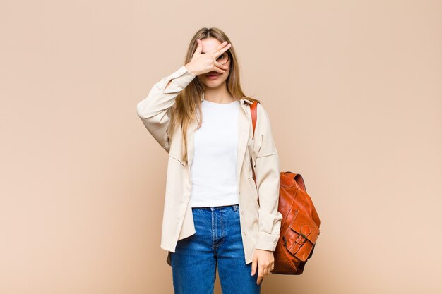 Blonde pretty woman looking shocked, scared or terrified, covering face with hand and peeking between fingers