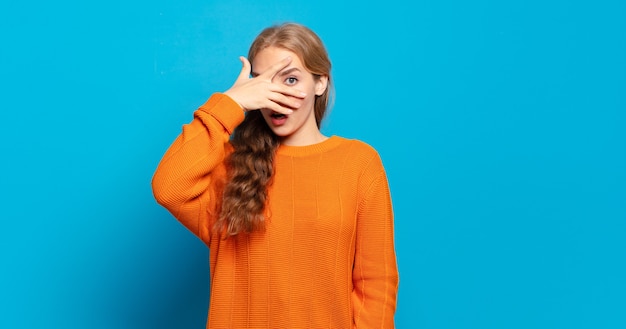 Blonde pretty woman looking shocked, scared or terrified, covering face with hand and peeking between fingers