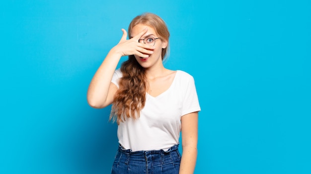 Blonde pretty woman looking shocked, scared or terrified, covering face with hand and peeking between fingers