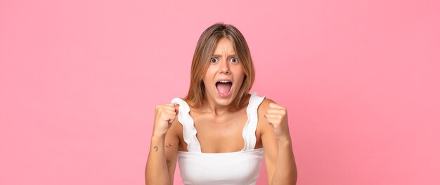 Bella donna bionda che grida in modo aggressivo con sguardo infastidito, frustrato, arrabbiato e pugni stretti, sentendosi furiosa