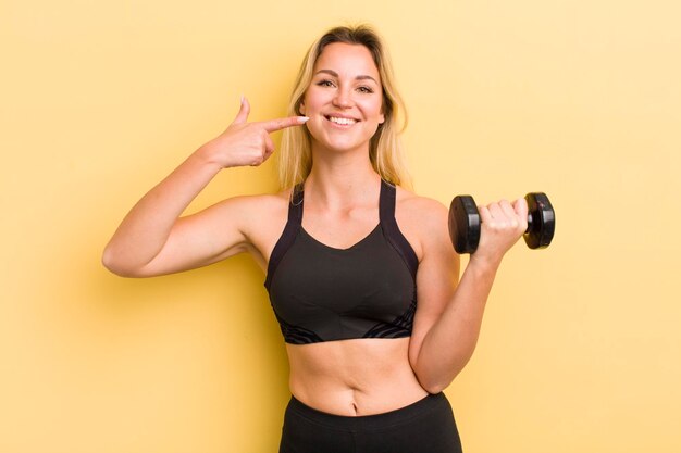 Blonde pretty woman smiling confidently pointing to own broad smile