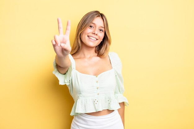 Blonde pretty woman smiling and looking happy, carefree and positive, gesturing victory or peace with one hand