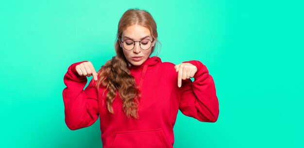 Blonde pretty woman with open mouth pointing downwards with both hands, looking shocked, amazed and surprised