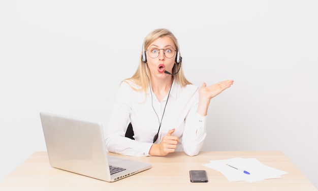 Bionda bella donna giovane donna bionda che sembra sorpresa e scioccata, con la mascella caduta tenendo un oggetto e lavorando con un laptop a