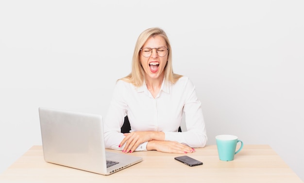 Blonde pretty woman young blonde woman shouting aggressively, looking very angry and working with a laptop