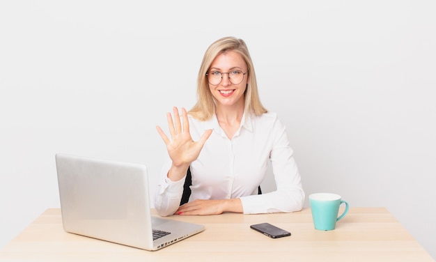 Blonde pretty woman young blonde woman smiling and looking friendly, showing number five and working with a laptop