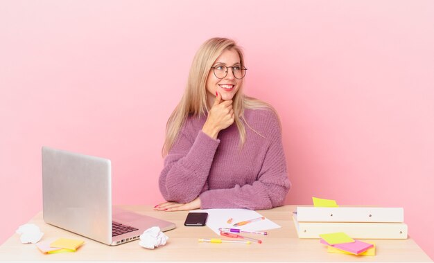 Blonde pretty woman young blonde woman smiling with a happy, confident expression with hand on chin and working with a laptop