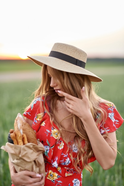 Photo a blonde in a red dress and a straw hat with french bread in her hands on a wheat field at sunset