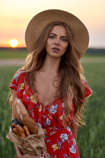 A blonde in a red dress and a straw hat with French bread in her hands on a wheat field at sunset