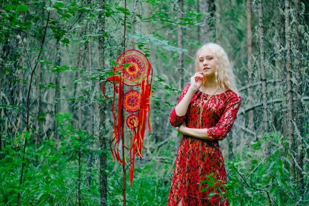 Blonde in a red dress in the woods next to a dreamcatcher