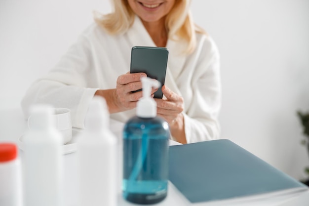 Blonde rijpe vrouw zit in cosmetologie centrum en maakt gebruik van mobiele telefoon surfen op het net