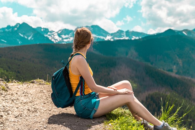 Blonde schattig meisje met een rugzak en een bril zit op een berg en geniet van de prachtige heuvels van de bergen op een zonnige dag.