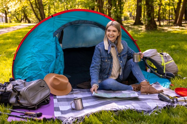 Blonde schattige vrouwelijke reiziger zit bij de tent in het bos
