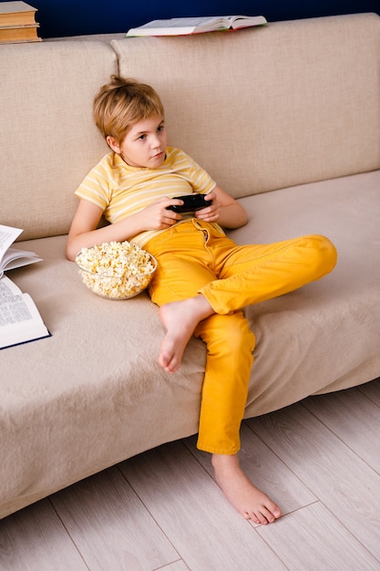 Blonde schoolboy boy plays video games, holds a gamepad, eats popcorn