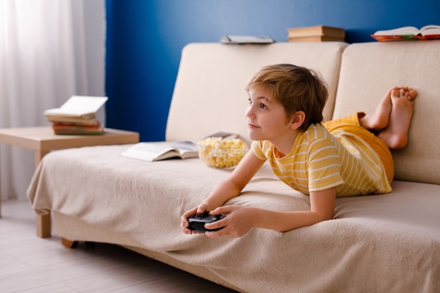 Blonde schoolboy boy plays video games, holds a gamepad, eats popcorn