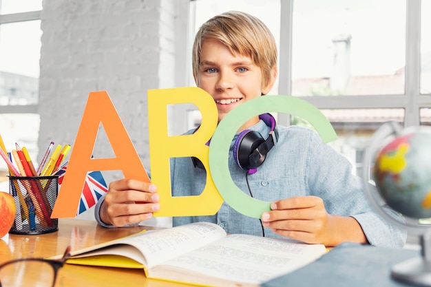 Blonde schooljongen die kleurrijke Engelse letters vasthoudt terwijl hij in het lichte klaslokaal zit