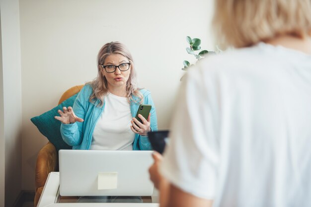 Blonde senior vrouw met bril met een telefoon legt iets uit aan een klant met behulp van een laptop en thuis werken