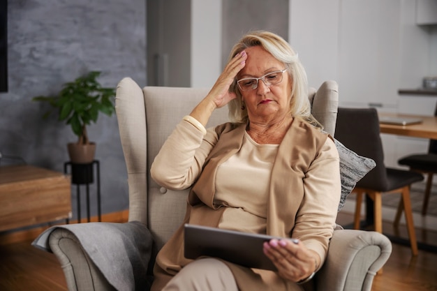 Blonde senior vrouw om thuis te zitten, met hoofdpijn en tablet te kijken om online adviezen te krijgen.