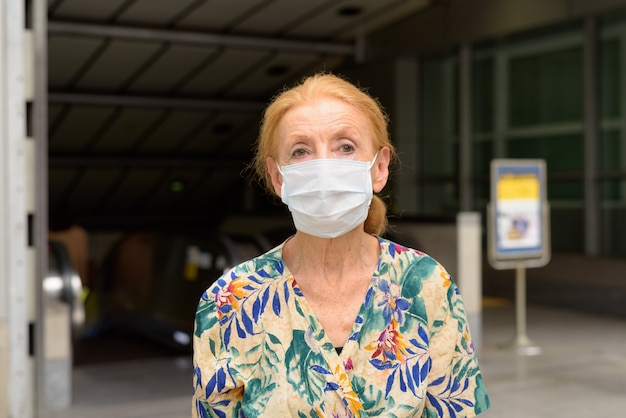 屋外の地下鉄の駅でコロナウイルスの発生からの保護のためのマスクを着ている金髪の年配の女性