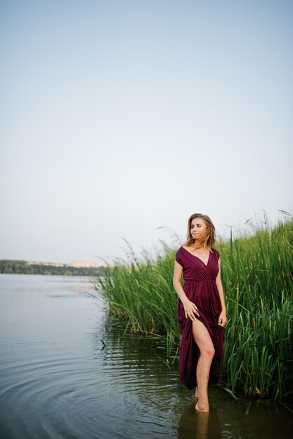 Blonde sensual woman in red marsala dress standing in water of lake with reeds