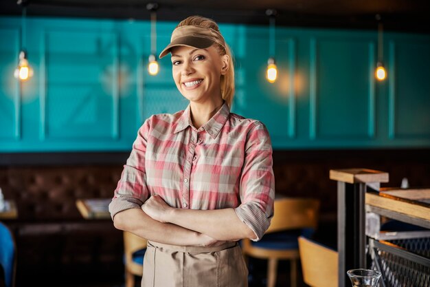 The blonde server wears a plaid shirt, cap and apron. She has her arms crossed and is looking straight at the front with a smile