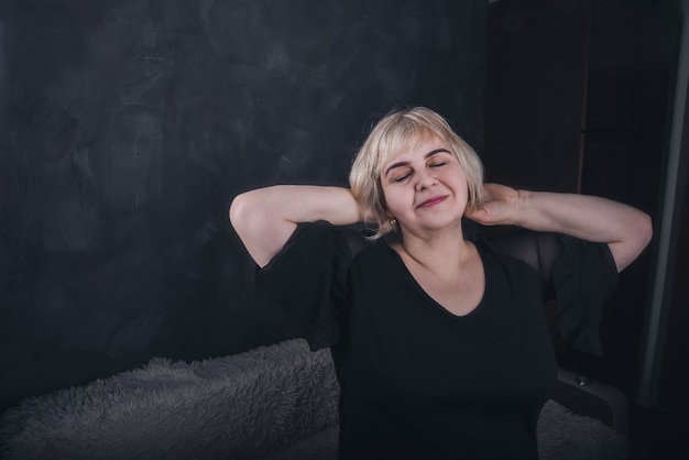 A blonde, short-haired, chubby older woman in black clothes on a solid background of a dark gray concrete.