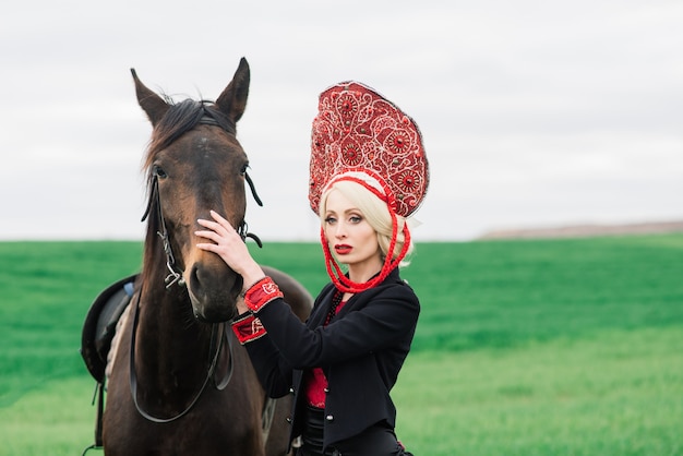 Блондинка славянская женщина в черном платье и головном уборе кокошник на поле с черной лошадью на закате