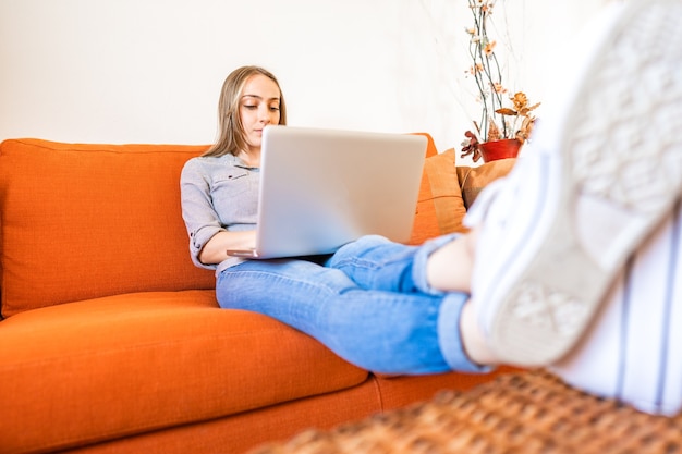 Blonde slimme gen z meisje met behulp van laptop om thuis te zitten bank om online te winkelen. jonge student geniet van internet om verbinding te maken met schoolnetwerk en klasgenoten. nieuwe normale activiteit van online lessen op afstand