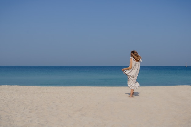 A blonde smiling girl in a maxi linen dress with fluttering hair jumping and dancing on a sand beach against blue sky and sea background