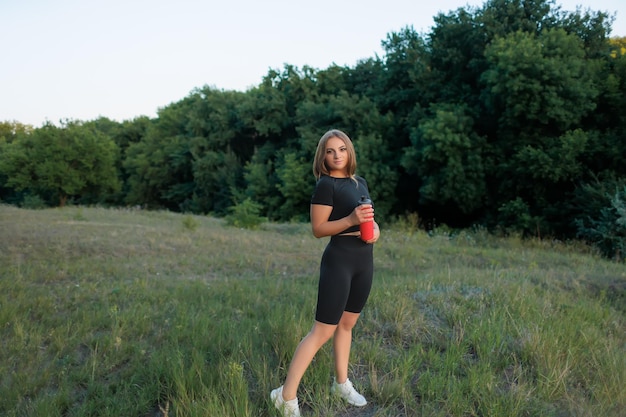 Blonde sportvrouw in het park poseren met een rode fles water bij zonsondergang