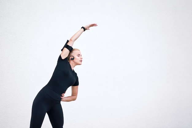 Blonde sporty girl wearing black sportswear listening to music and stretching on white wall. Concept active and healthy life.